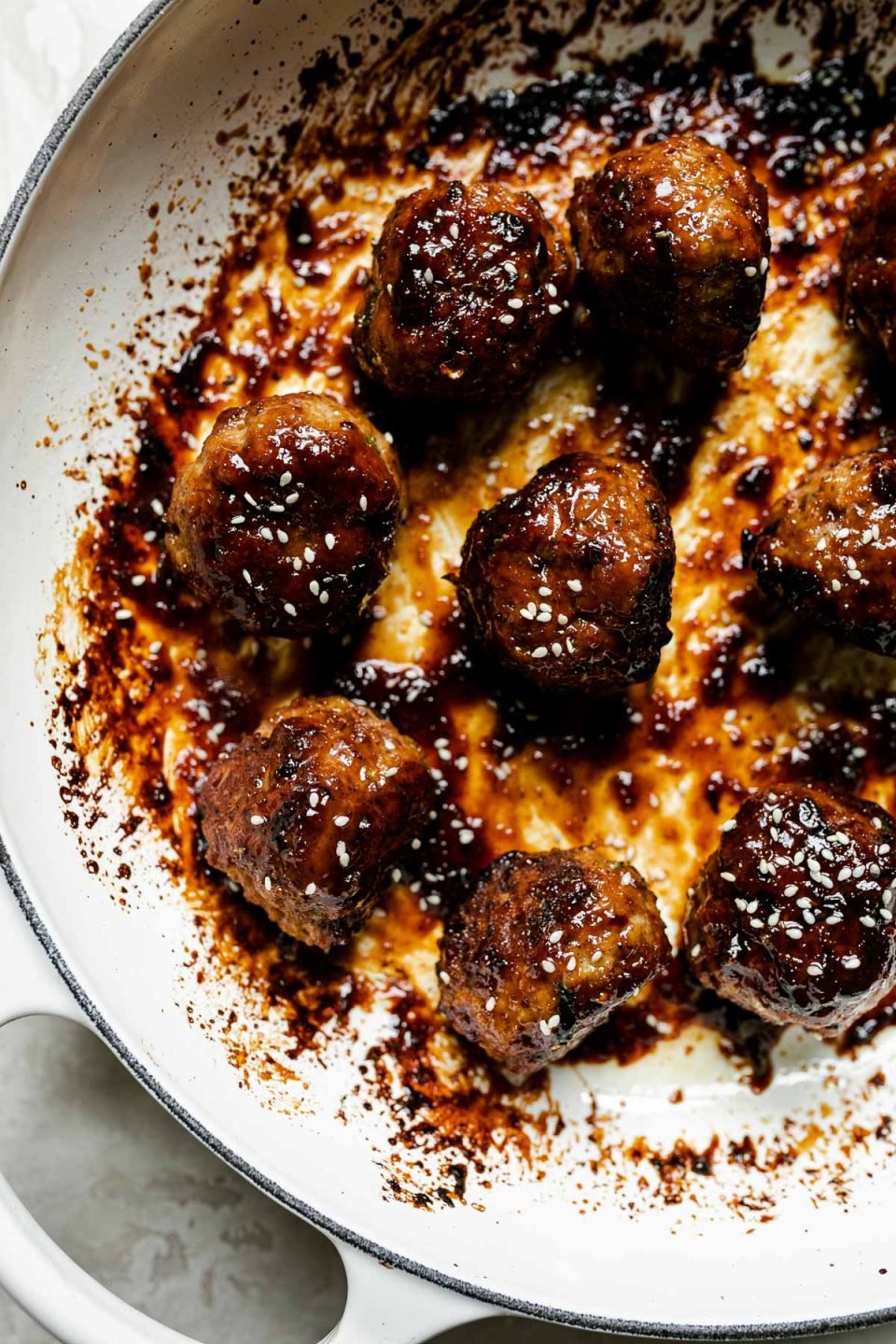 Close up of glazed meatballs in white enameled braising pan atop a creamy cement surface. The meatballs are garnished with sesame seeds.