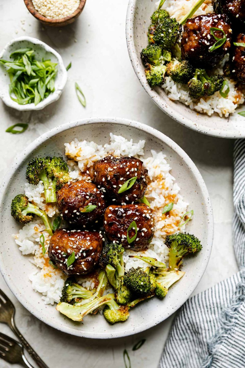 Soy-glazed ginger turkey meatballs plated in large ceramic bowls over rice with roasted broccoli. The meatballs are garnished with sesame seeds & sliced green onions. The bowls sit atop a creamy cement surface surrounded by a gray striped linen napkin, forks, a small wooden bowl filled with sesame seeds & a small ceramic bowl of sliced green onions.