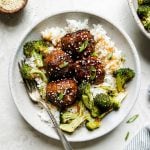 Soy-glazed ginger turkey meatballs plated in large ceramic bowls over rice with roasted broccoli. The meatballs are garnished with sesame seeds & sliced green onions. The bowls sit atop a creamy cement surface surrounded by a gray striped linen napkin, forks, a small wooden bowl filled with sesame seeds & a small ceramic bowl of sliced green onions.