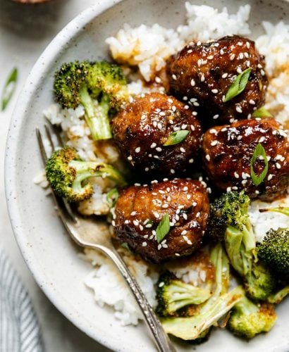Soy-glazed ginger meatballs plated in large ceramic bowl over rice with roasted broccoli. The meatballs are garnished with sesame seeds & sliced green onions, & a fork is nestled into the rice. The bowl sits atop a creamy cement surface, surrounded by a striped linen napkin & a small wooden bowl of sesame seeds.