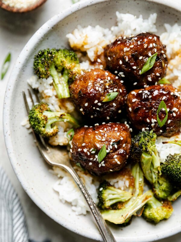 Soy-glazed ginger meatballs plated in large ceramic bowl over rice with roasted broccoli. The meatballs are garnished with sesame seeds & sliced green onions, & a fork is nestled into the rice. The bowl sits atop a creamy cement surface, surrounded by a striped linen napkin & a small wooden bowl of sesame seeds.