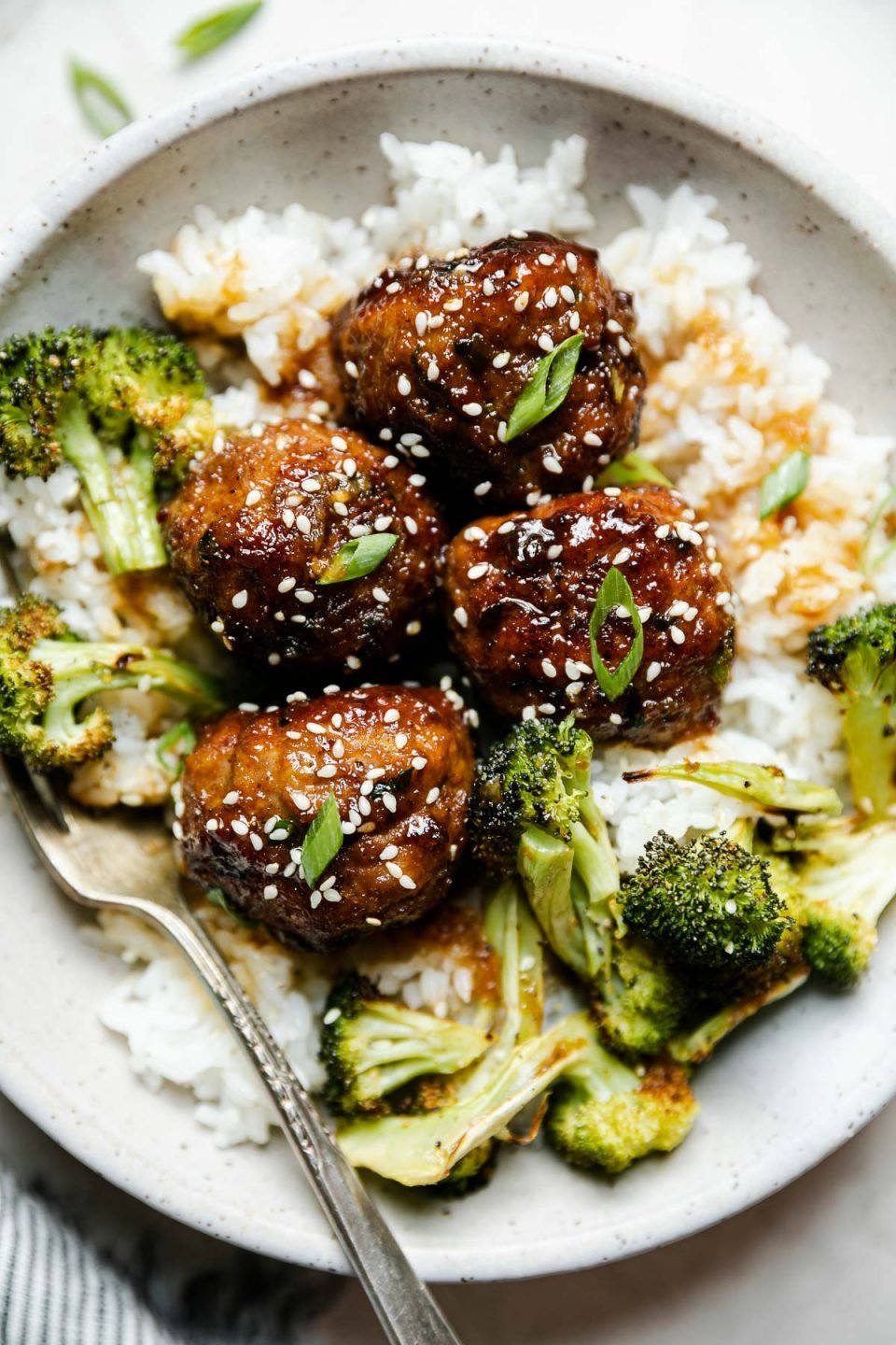 Soy-glazed ginger meatballs plated in large ceramic bowl over rice with roasted broccoli. The meatballs are garnished with sesame seeds & sliced green onions, & a fork is nestled into the rice. The bowl sits atop a creamy cement surface, surrounded by a striped linen napkin & a small wooden bowl of sesame seeds.