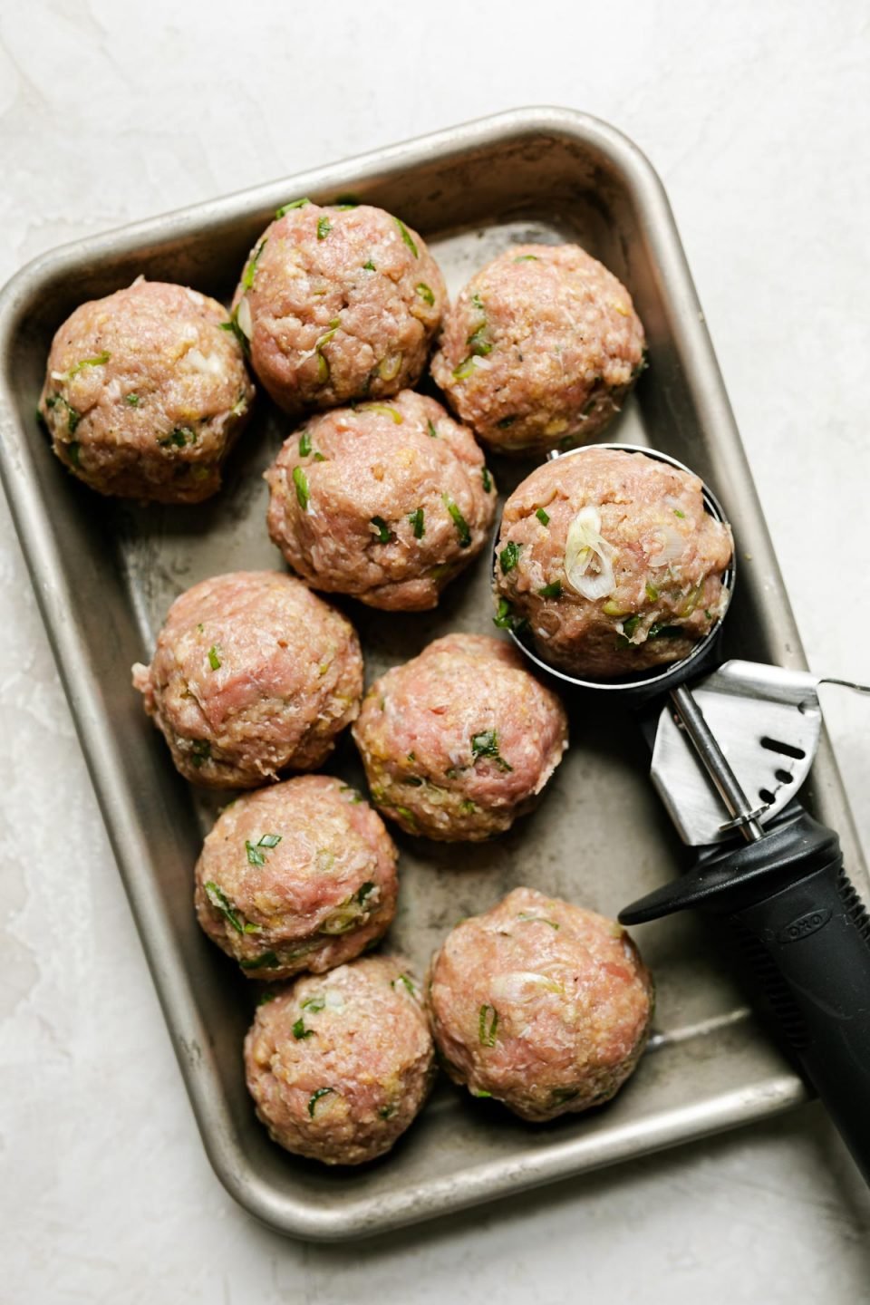 Rolling out ginger meatballs – 9 large turkey meatballs on a small baking sheet with a 10th meatball in a scoop alongside them. The baking sheet sits atop a creamy cement surface.