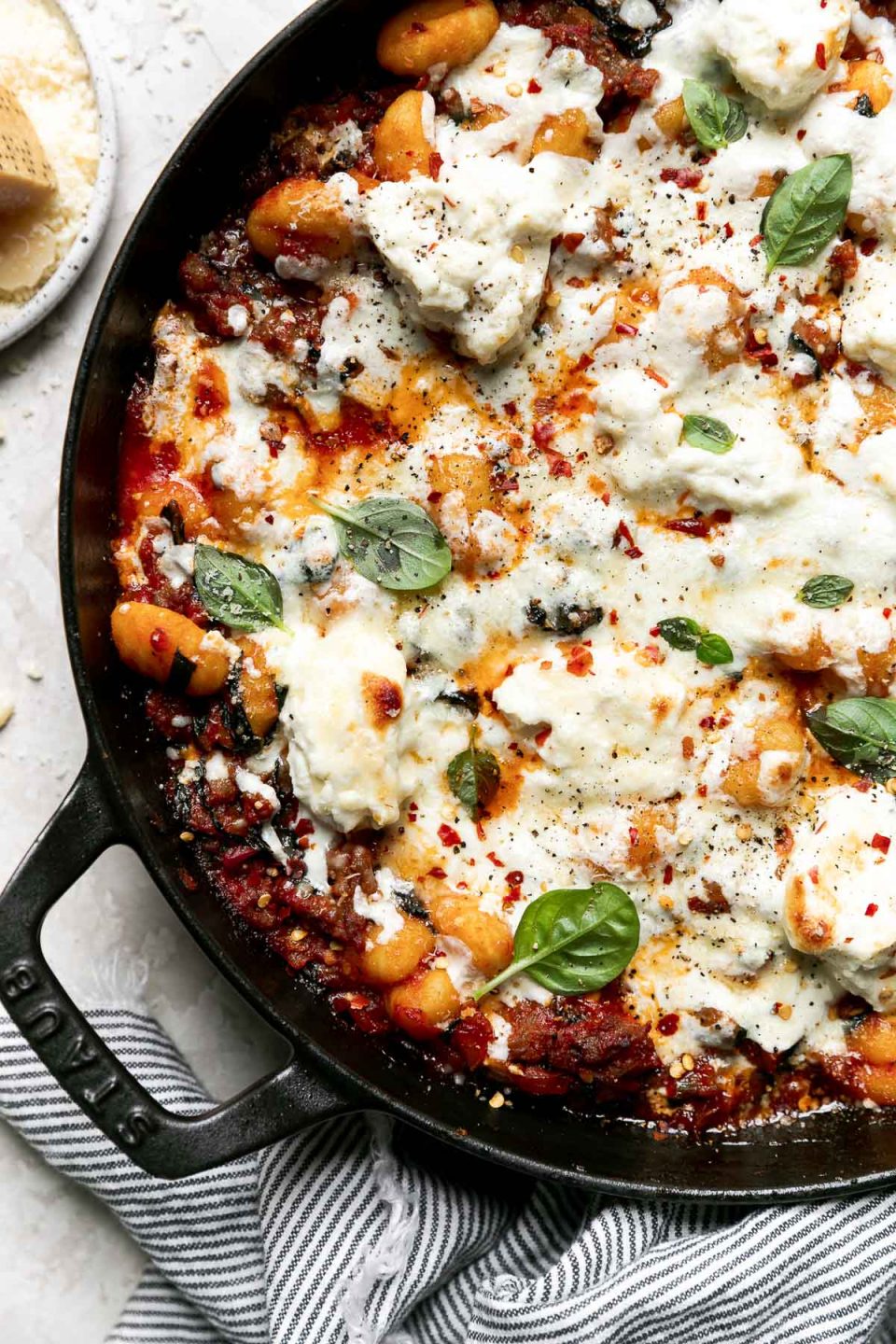 Overhead shot of baked gnocchi in a large oven-safe skillet, atop a creamy white surface. The pasta bake is garnished with fresh basil & ground black pepper. A blue & white striped linen napkin and a rind of freshly grated Parmigiano Reggiano cheese rests on the surface alongside the skillet.