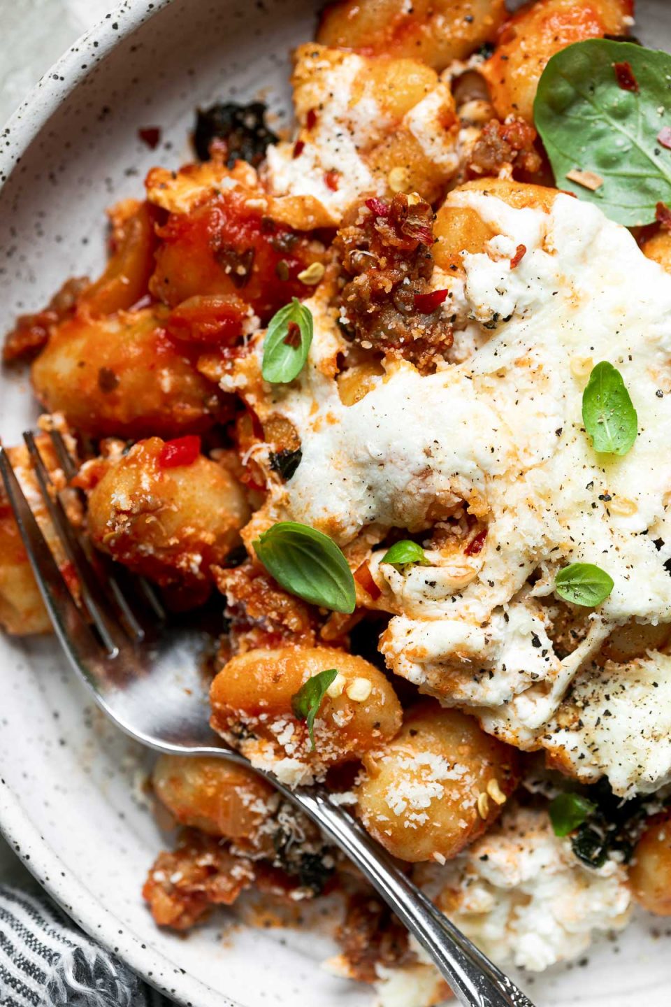 A close up of baked gnocchi shown on a speckled plate, topped with fresh basil & ground black pepper. A silver fork rests on the plate and a blue & white striped linen napkin rests alongside the plate.