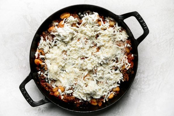 Overhead shot of browned Italian sausage, peppers, onions, & kale covered in dots of fresh ricotta, mozzarella, & a sprinkle of fresh parmesan for oven baked gnocchi in a large oven-safe skillet, atop a creamy white surface