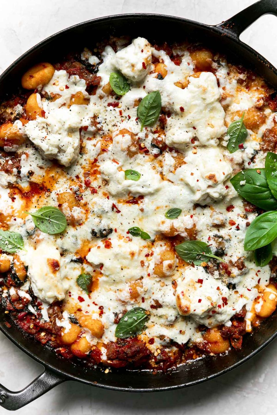 Overhead shot of baked gnocchi in a large oven-safe skillet, atop a creamy white surface. The pasta bake is garnished with fresh basil & ground black pepper.