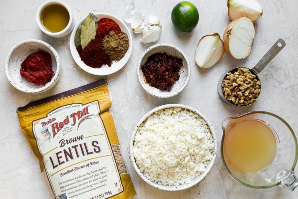 Cauliflower Lentil Tacos ingredients arranged on a white cement surface: chopped walnuts, olive oil, yellow onion, cauliflower rice, garlic, chipotle pepper, tomato paste, chili powder, ground cumin, smoked paprika, bay leaf, Bob's Red Mill Brown Lentils, vegetable stock, and lime.