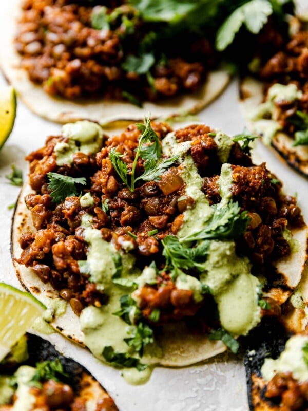 An angled close up shot of Cauliflower Lentil Tacos arranged on a white cement surface, topped with cilantro lime crema sauce & chopped cilantro. There are a few lime wedges surrounding the tacos.