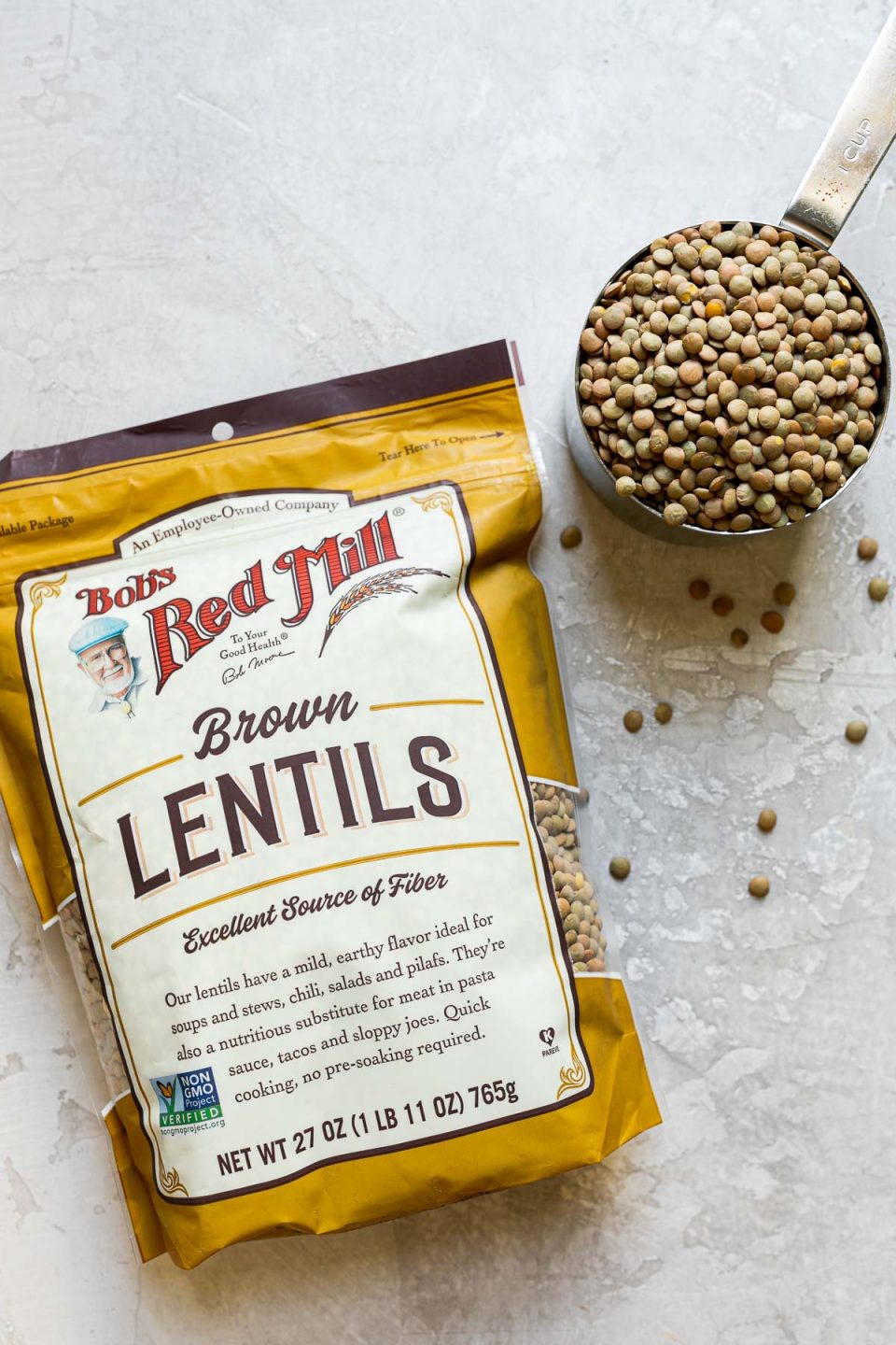 A single package of Bob's Red Mill Brown Lentils lays flat on a white cement surface. A 1 cup dry measuring cup filled with dry brown lentils sits alongside the package and the surface has a sprinkling of dry brown lentils scattered between the package & the measuring cup.