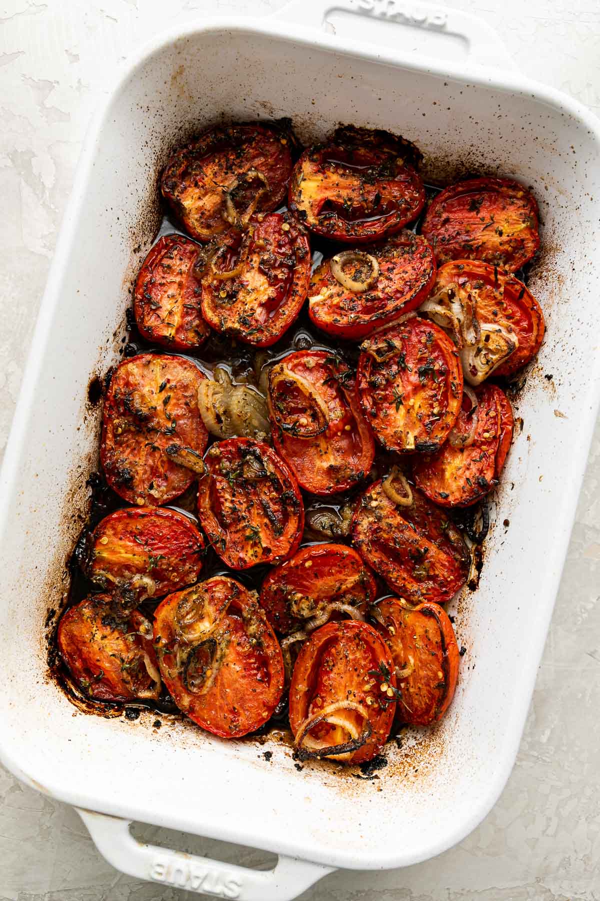 Oven roasted roma tomatoes, shallots, & garlic in a white Staub baking dish. The dish sits atop a creamy white surface.
