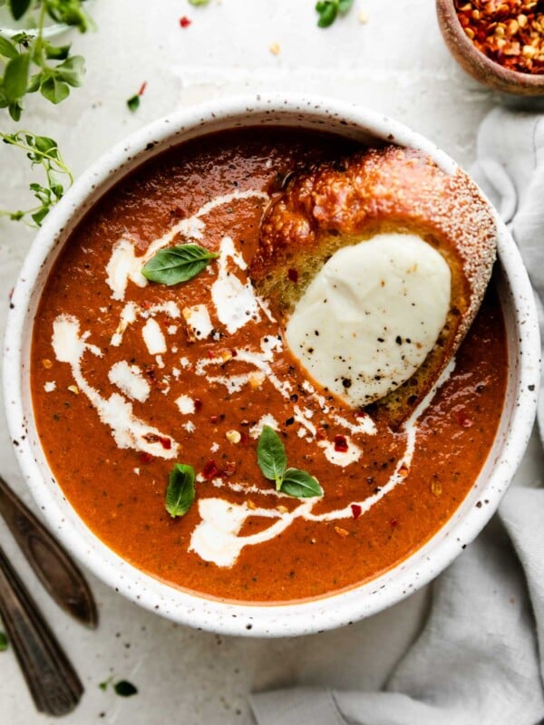 Oven Roasted Tomato soup, drizzled with heavy cream & topped with crushed red pepper flakes & a few fresh basil leaves, served in a speckled ceramic bowl. The bowl sits atop a creamy white surface and is surrounded by a jar of fresh herbs, silverware, a small wooden pinch bowl filled with crushed red pepper flakes, and a light gray linen napkin. Inside of the bowl of soup rests a fresh mozzarella crostini.