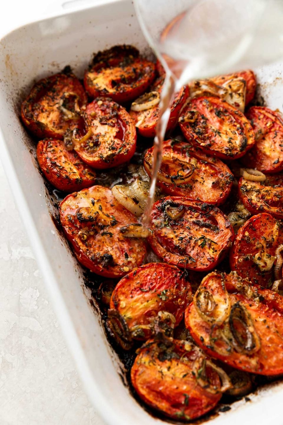 Oven roasted roma tomatoes, shallots, & garlic in a white Staub baking dish. The dish sits atop a creamy white surface. Vermouth is being poured from a clear glass measuring to deglaze the bottom of the baking dish to make roasted tomato basil soup.