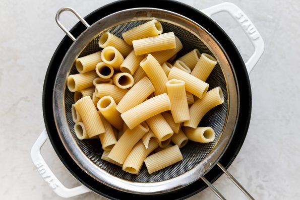 An overhead shot of a mesh strainer sitting over a large white pot filled with strained rigatoni pasta noodles. The pot sits atop a creamy white surface.