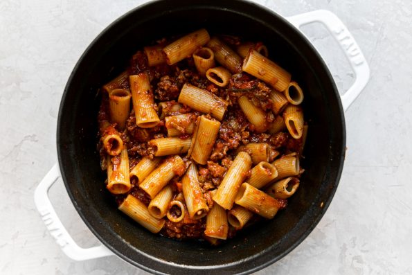 An overhead shot of a large pot filled with weeknight bolognese sauce mixed with rigatoni pasta noodles. The pot sits atop a creamy white surface.