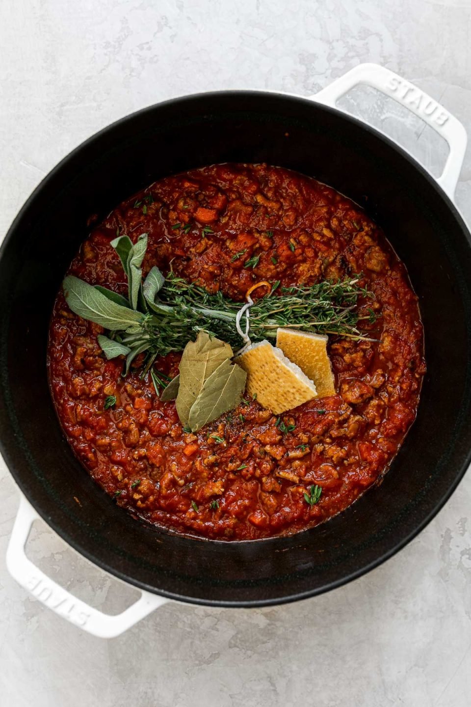 Weeknight bolognese sauce made with turkey in a large pot sits atop a creamy white surface. A tied bunch of fresh herbs, two dried basil leaves, & a parmesan rind have been added atop of the sauce to build flavor.