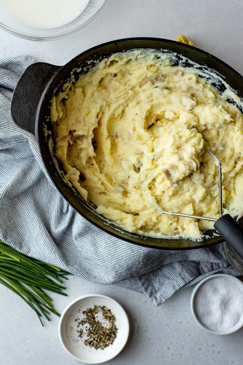 An overhead shot of a dutch oven filled with Roasted Garlic Buttermilk Mashed Potatoes. The dutch oven sits atop a creamy white textured surface. A potato masher rests inside the dutch oven. A blue and white striped linen napkin, a pile of chive scapes, two white pinch bowls filled with ground black pepper and kosher salt, and a clear glass bowl filled with buttermilk ​also rest on the surface & surround the serving bowl.