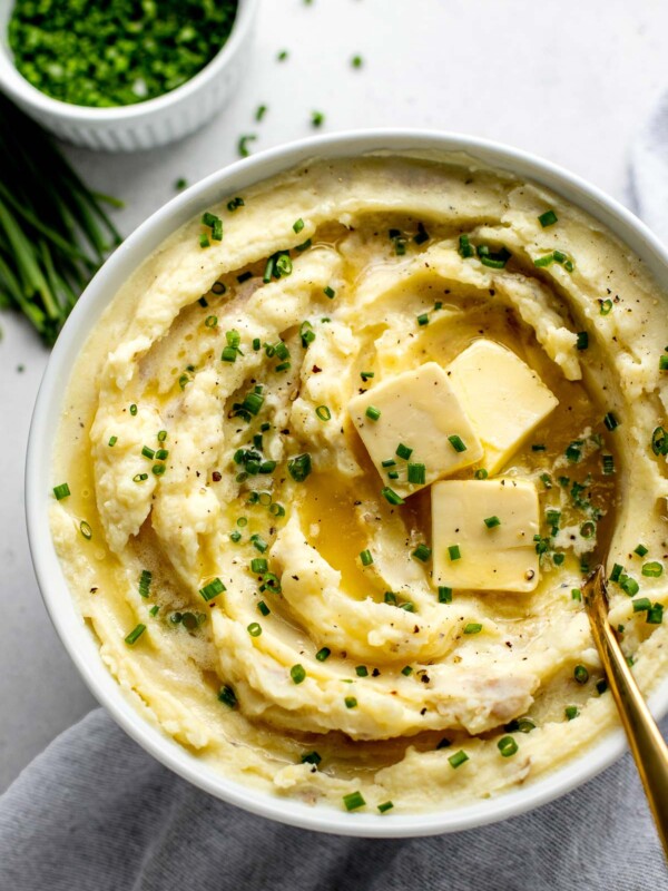 An overhead shot of a white serving bowl filled with Roasted Garlic Buttermilk Mashed Potatoes. The bowl sits atop a creamy white textured surface. A gold serving spoon rests inside the mashed potatoes and three pads of sliced butter sit atop the potatoes in a pool of melted butter. The potatoes are garnished with black pepper and snipped chives. A light gray linen napkin, a white ramekin filled with snipped chives, and a pile of chive scapes and loose snipped chives also rest on the surface & surround the serving bowl.