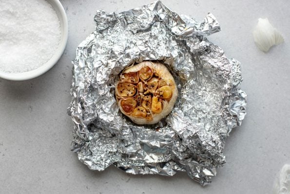 An overhead shot of a bulb of roasted garlic that sits atop a crumpled sheet of aluminum foil previously used to roast it. A small white pinch bowl filled with kosher salt and loose outer garlic skin pieces rest alongside the roasted garlic bulb.