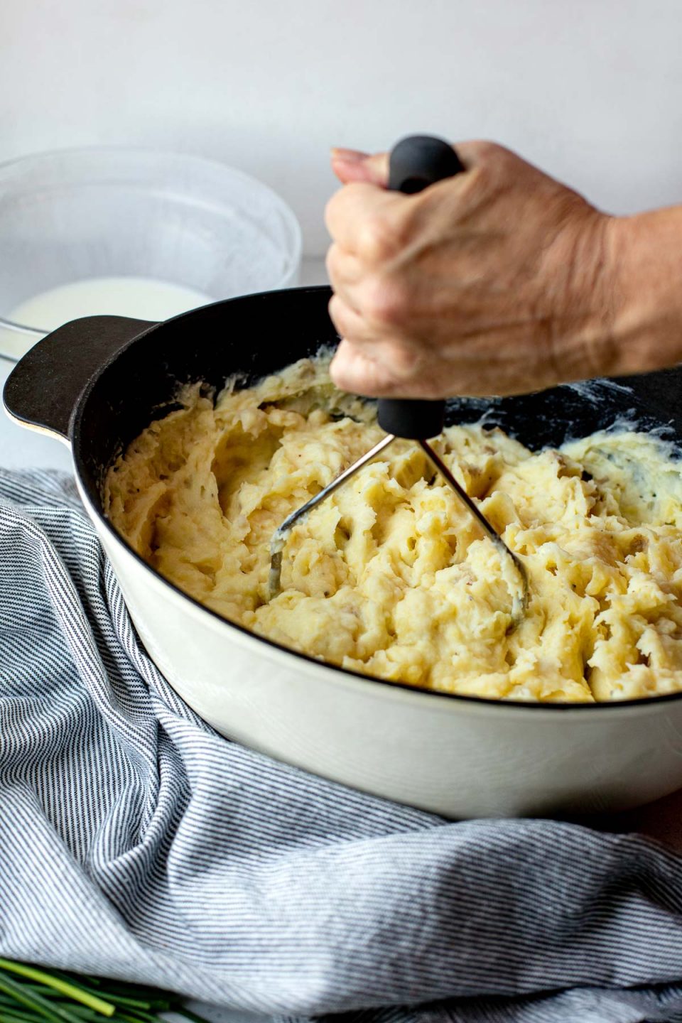 A side angle shot of a white dutch oven filled with Roasted Garlic Buttermilk Mashed Potatoes. The dutch oven sits atop a creamy white textured surface. A woman's hand holds a potato masher and is using it to mash up the potatoes within the dutch oven. A blue and white striped linen napkin, a pile of chive scapes, and a clear glass bowl filled with buttermilk ​also rest on the surface & surround the serving bowl.