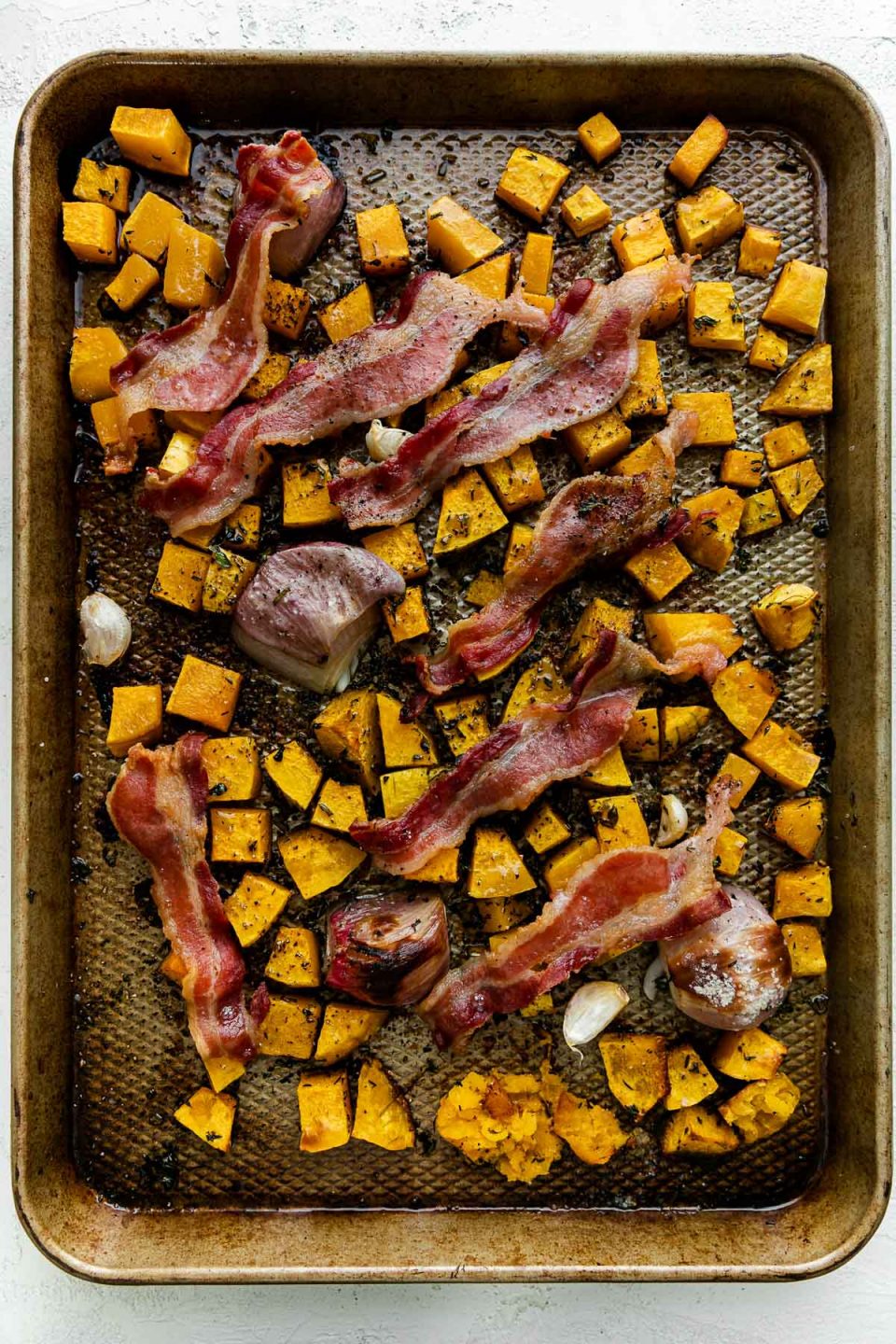 Cubed butternut squash, shallots, & garlic seasoned with olive oil & fresh herbs with bacon draped over top on a baking sheet after roasting. The baking sheet sits atop a white textured surface.