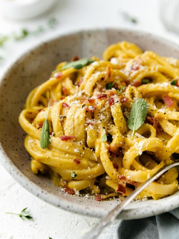 Creamy Butternut Squash Pasta shown in speckled ceramic pasta bowl, topped with crispy bacon, grated parmesan, ground black pepper, & fresh sage leaves. A silver fork rests inside of the pasta bowl & the bowl sits atop a white textured surface surrounded by a light blue linen napkin, & a small white bowl filled with freshly grated parmesan.