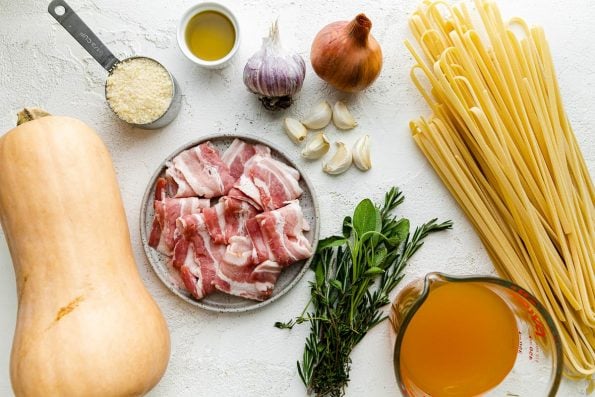 Butternut squash pasta ingredients arranged on a light plaster surface: butternut squash, shallots, garlic, olive oil, fresh herbs, bacon, vegetable stock, grated parmesan, linguine noodles.