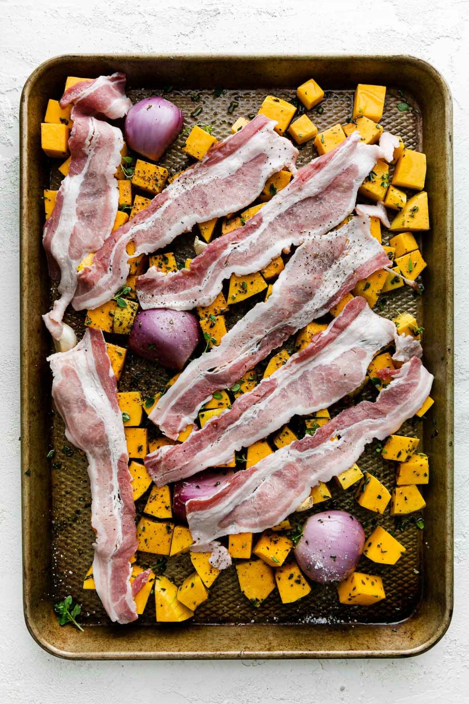 Cubed butternut squash, shallots, & garlic seasoned with olive oil & fresh herbs with bacon draped over top on a baking sheet before roasting. The baking sheet sits atop a white textured surface.