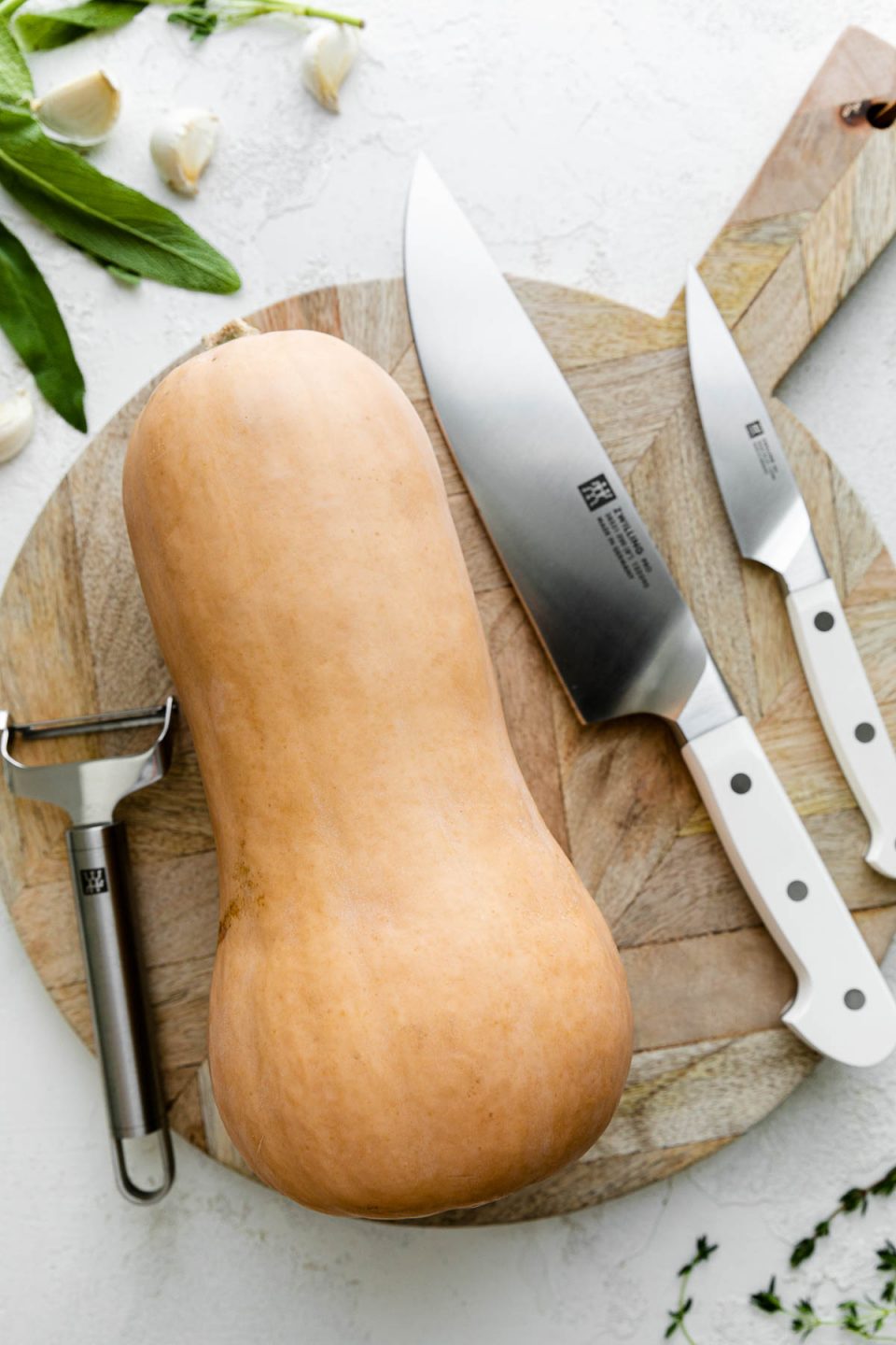 A whole butternut squash rests on top of a round wooden cutting board alonside Zwilling Pro Le Blanc Knives & a Zwilling Pro Y-Peeler. The cutting board sits atop a white textured surface while fresh herbs and cloves of garlic surround the cutting board.