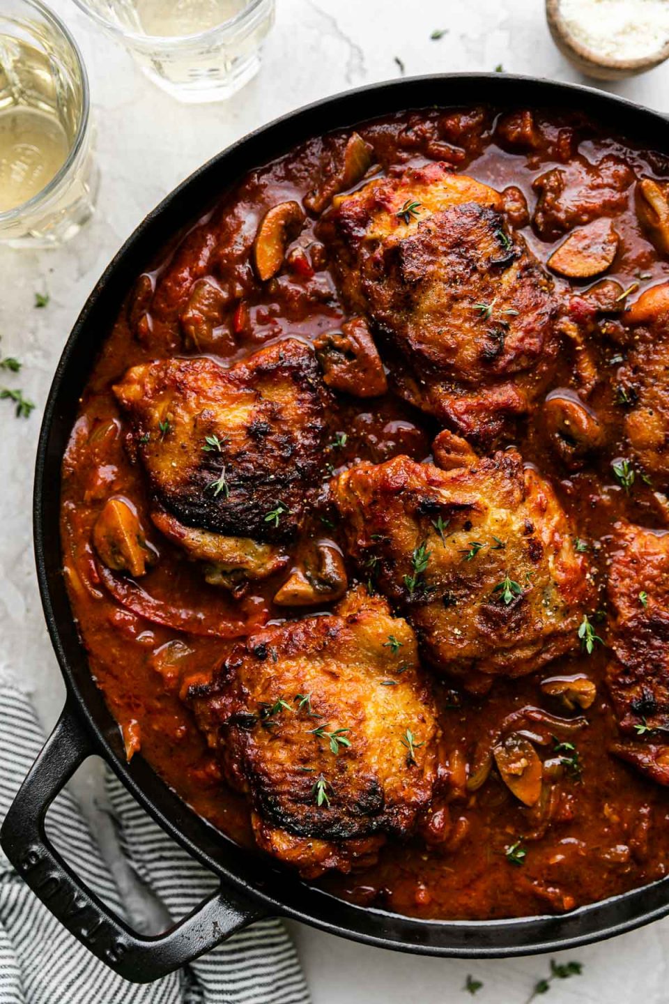 A heavy-bottomed cast iron skillet filled with chicken cacciatore sits atop a creamy white plaster surface. Two glasses of white wine, a small wooden pinch bowl filled with grated parmesan, a white and blue striped linen napkin, and a few sprigs of fresh thyme surround the skillet.