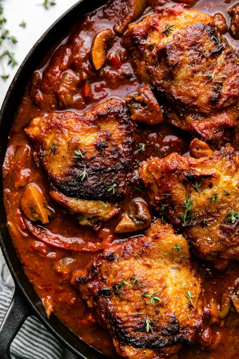 A close up of chicken cacciatore in a black heavy-bottomed cast iron skillet. The skillet sits atop a creamy white plaster surface with a blue and white striped napkin resting alongside and a few sprigs of thyme also sprinkled next to the skillet.
