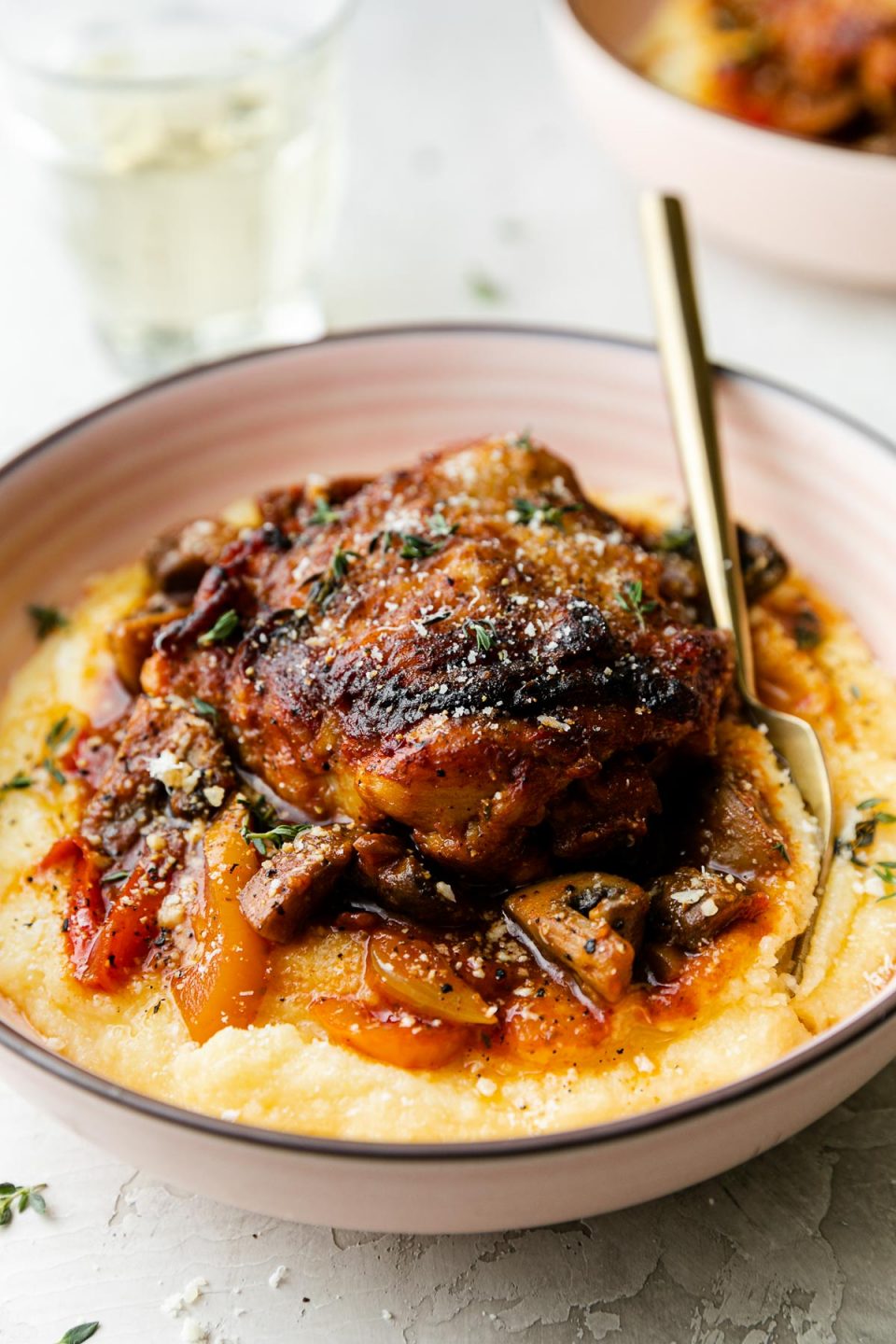 A side angle shot of chicken cacciatore served over creamy polenta in a pink bowl. A gold fork is tucked inside of the bowl. The bowl sits atop a creamy white plaster surface and a few sprigs of fresh thyme surround the bowl. Another bowl of chicken cacciatore and a single glass of white wine rest in the background.