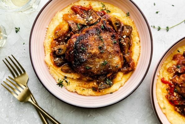 Two pink ceramic bowls filled with chicken cacciatore served over creamy polenta sit atop a creamy white plaster surface. A few sprigs of fresh thyme, two gold forks, and two glasses of white wine surround the bowl.
