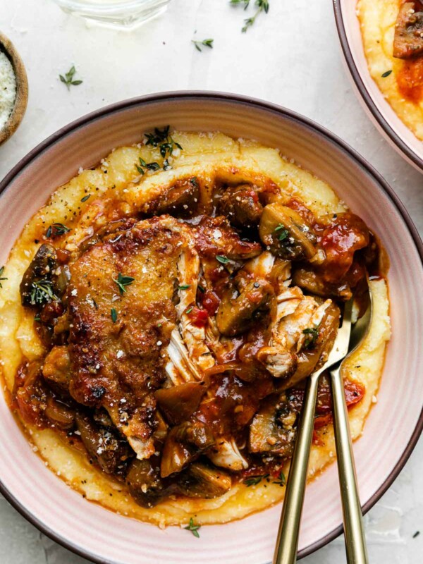 Two pink ceramic bowls filled with chicken cacciatore served over creamy polenta sit atop a creamy white plaster surface. A few sprigs of fresh thyme, a small wooden pinch bowl filled with grated parmesan cheese, and a single glass of white wine rest near the bowl. A gold fork and spoon rest inside of the bowl that is centered in the frame.