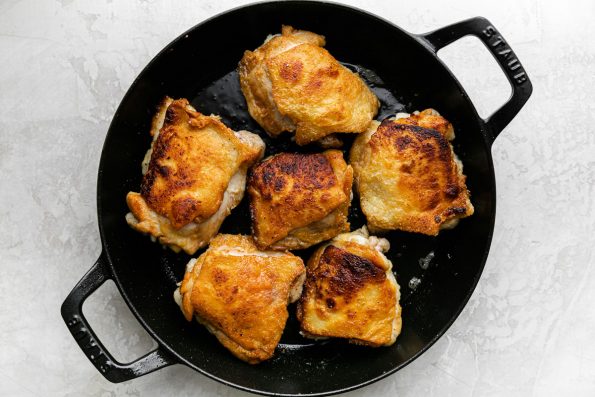 How to Make Chicken Cacciatore, Step 2: Chicken thighs brown in an oiled cast iron skillet until golden brown and rendered. The cast iron skillet rests atop a creamy white plaster surface.