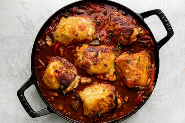 How to Make Chicken Cacciatore, step 6: Browned chicken thighs are nestled into a cacciatore sauce inside of a heavy-bottomed cast iron skillet. The skillet rests atop a creamy white plaster surface.