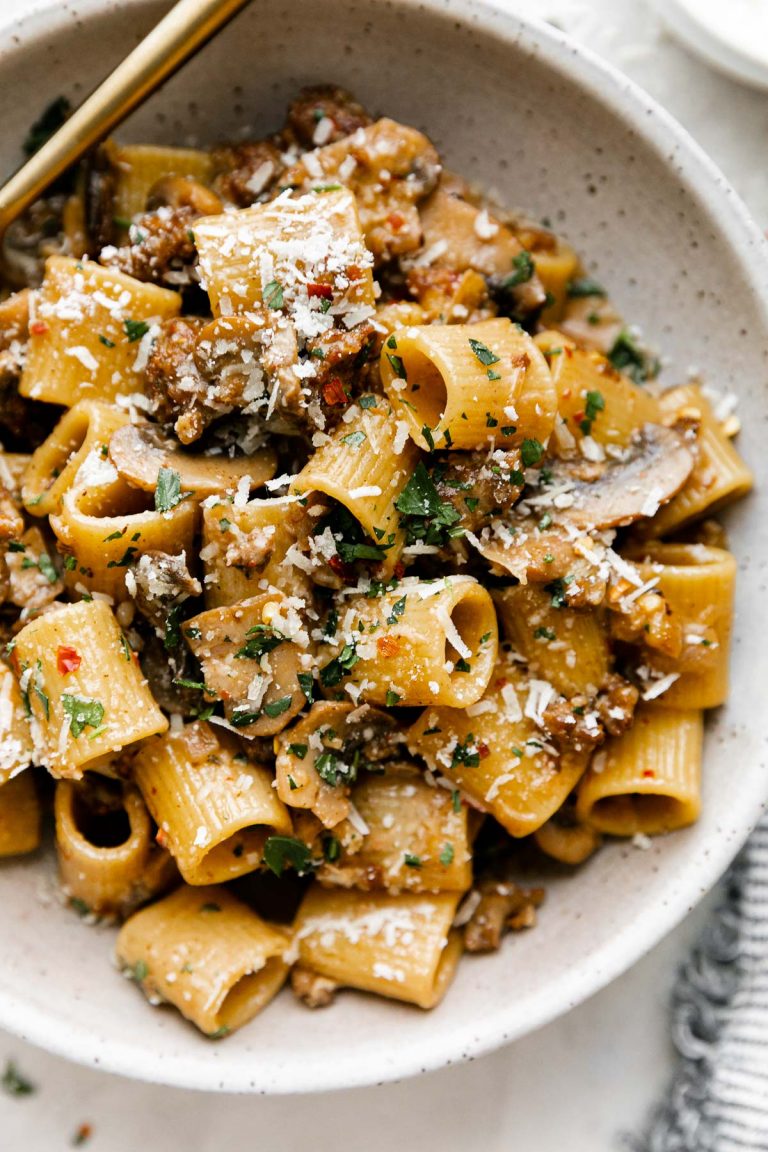 A close up shot of Creamy Marsala Pasta with Italian Sausage and Mushrooms inside of a white speckled ceramic bowl sits atop a creamy white plaster surface. A gold fork rests inside of the bowl and a blue and white striped napkin rests alongside the bowl.