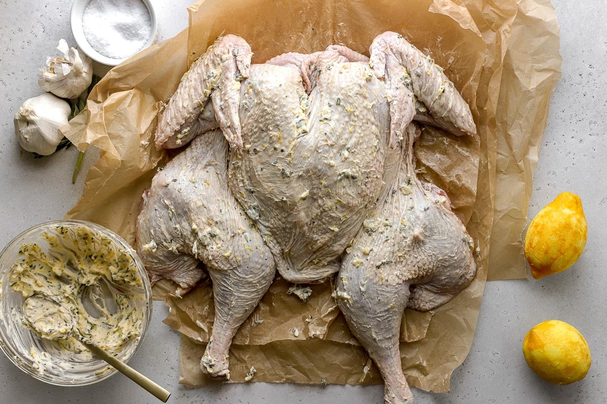 A spatchcocked turkey seasoned with lemon herb butter directly on the exterior skin rests breast side up on a few sheets of brown parchment paper. A bowl of mixed lemon herb butter with a mixing spoon resting inside, a few bulbs of garlic, loose herbs, two zested lemons, and a white ramekin filled with kosher salt rest alongside the turkey. All items sit atop a lightly colored textured surface.