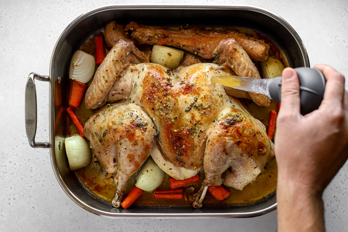 A spatchcocked turkey seasoned with lemon herb butter is placed within a roasting pan surrounded by garlic cloves, yellow onion, carrots, and the turkey neck and spine. A man's hand holds a turkey baster to baste the surface of the turkey with drippings from the bottom of the pan. The roasting pan sits atop ​a lightly colored textured surface.