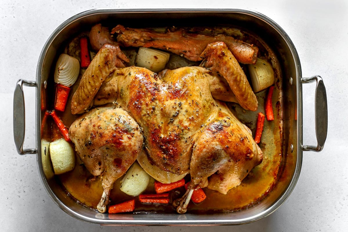 An overhead shot of a roasted spatchcocked turkey seasoned with lemon herb butter placed within a roasting pan surrounded by garlic cloves, yellow onion, carrots, and the turkey neck and spine. Turkey drippings have formed at the bottom of the roasting pan and the pan itself sits atop ​a lightly colored textured surface.