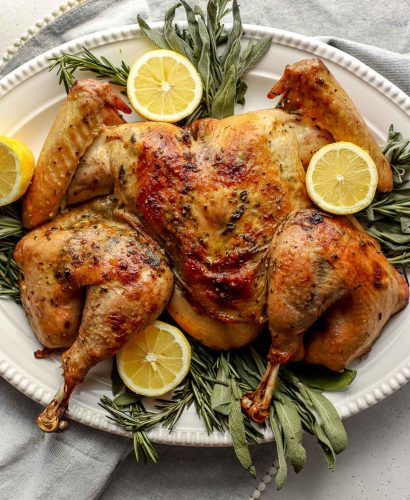 An overhead shot of a roasted spatchcocked turkey seasoned with lemon herb butter arranged on a white serving platter. The platter has been garnished with various fresh herbs and lemon slices. A light gray linen napkin is tucked underneath the platter and the platter sits atop ​a lightly colored textured surface.