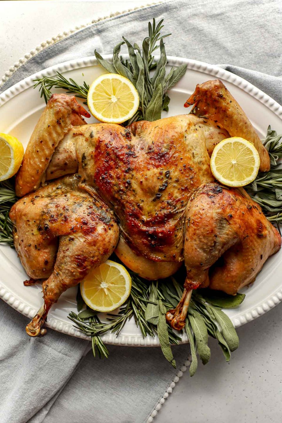An overhead shot of a roasted spatchcocked turkey seasoned with lemon herb butter arranged on a white serving platter. The platter has been garnished with various fresh herbs and lemon slices. A light gray linen napkin is tucked underneath the platter and the platter sits atop ​a lightly colored textured surface.