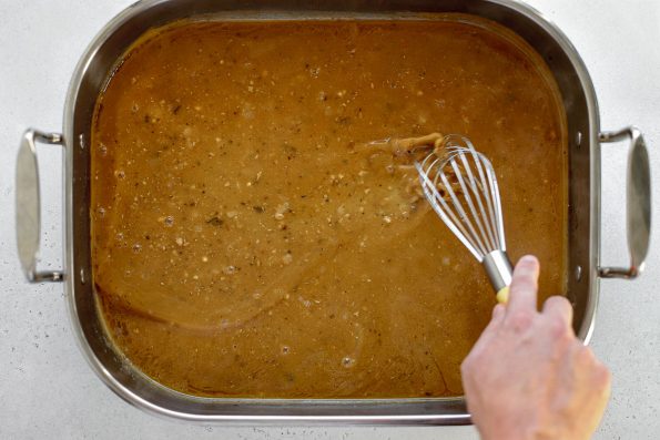 An overhead shot of a roasting pan filled with Easy Turkey Gravy. A person's hand holds a metal whisk that is being used to combine the gravy to help it reduce and thicken. The metal roasting pan sits atop a lightly colored textured surface.