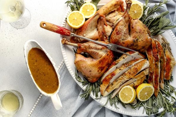 An overhead shot of a roasted spatchcocked turkey seasoned with lemon herb butter that has been carved and arranged on a white serving platter. The platter has been garnished with various fresh herbs and lemon slices. A silver carving fork with a wooden handle rests on the side of the platter. A white gravy boat filled with easy turkey gravy and two glasses of white wine sit alongside the platter. A light blue linen napkin lined with white pom fringe is tucked underneath the platter and all items sits atop ​a lightly colored textured surface.