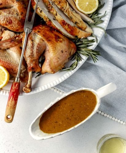 An overhead shot of a roasted spatchcocked turkey seasoned with lemon herb butter that has been carved and arranged on a white serving platter. The platter has been garnished with various fresh herbs and lemon slices. A silver carving fork with a wooden handle rests on the side of the platter. A white gravy boat filled with easy turkey gravy and a glass of white wine sit alongside the platter. A light blue linen napkin lined with white pom fringe is tucked underneath the platter and all items sits atop ​a lightly colored textured surface.
