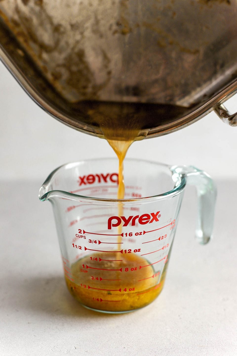 Roasted turkey pan drippings are being carefully poured into a liquid measuring cup from a roasting pan. The measuring cup sits atop a light colored textured surface.