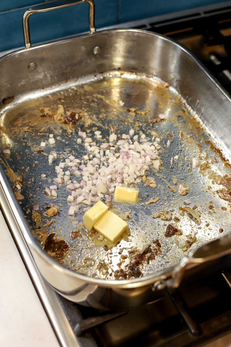 How to Make Easy Turkey Gravy, step 3: A roasting pan is placed over two burners on a stove top over low heat. Chopped shallots and unsalted butter are added to the roasting pan to cook along with some of the leftover turkey fat.