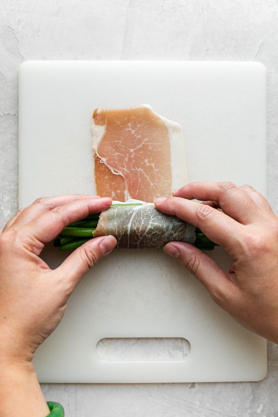A woman's hands work to slowly roll and wrap a pile of neatly arranged green beans, running in the same direction, with a single piece of prosciutto that is resting atop a white cutting board. The cutting board sits atop a creamy white plaster surface.