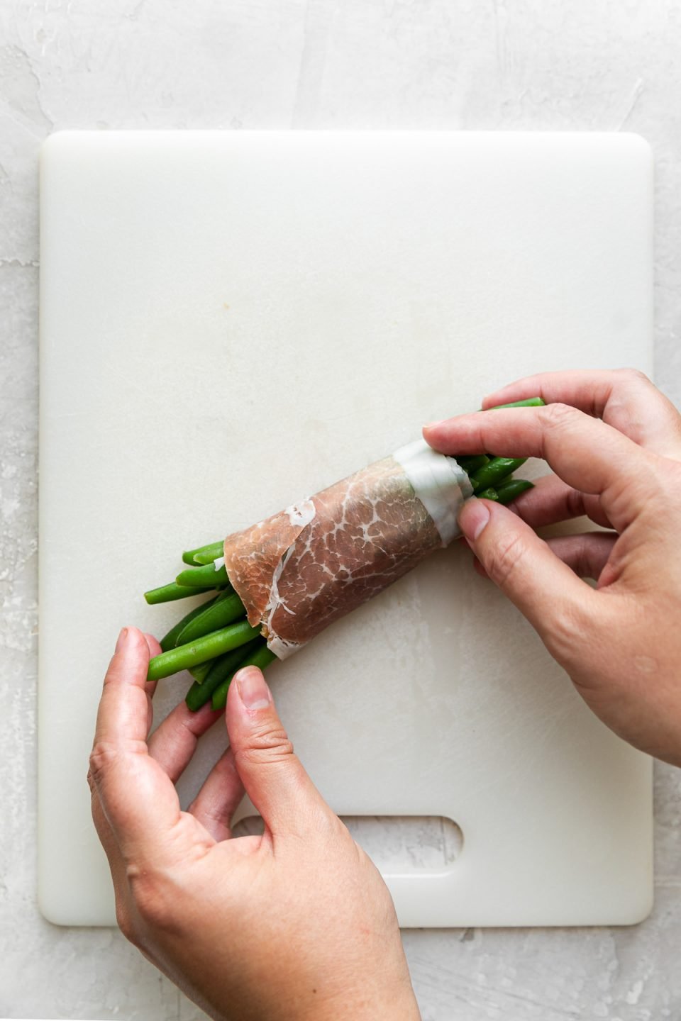 A woman's hands hold a finished prosciutto-wrapped green bean bundle that sits atop a white cutting board. The cutting board sits atop a creamy white plaster surface.