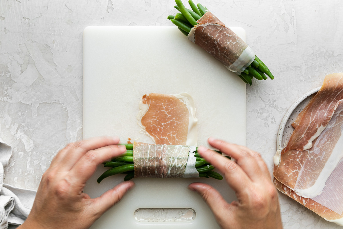 A woman's hands work to wrap a second pile of fresh green beans with prosciutto atop a white cutting board while a finished prosciutto-wrapped green bean bundle rests above it also atop the cutting board. To the right of the cutting board is a gray speckled ceramic plate holding sheets of prosciutto and to the left of the cutting board rests a light gray linen napkin. The cutting board, napkin, and plate all sit atop a creamy white plaster surface.